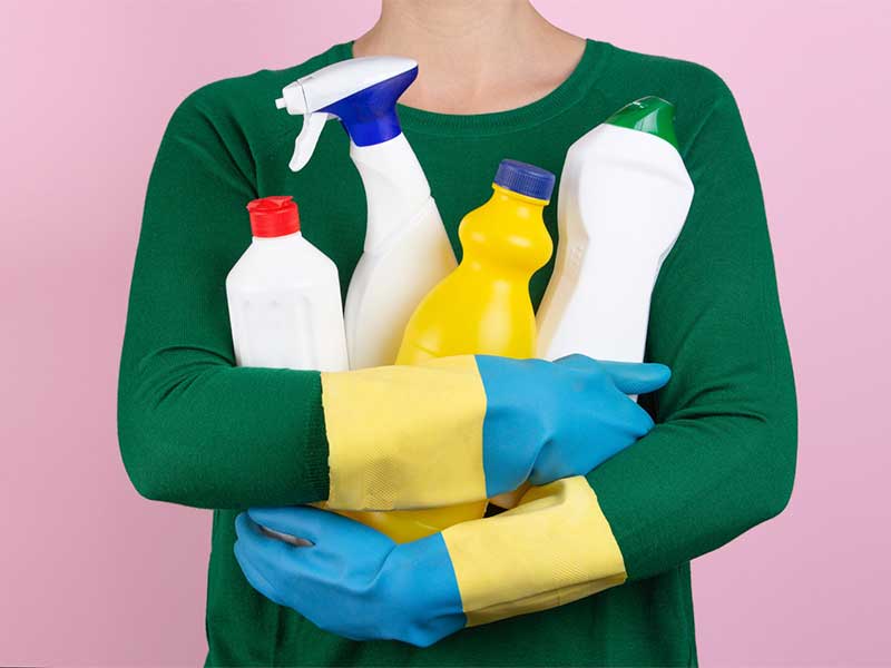 A imagem mostra o torso de uma mulher branca. Ela veste blusa verde e usa luvas azuis e amarelas nas mãos. Está segurando quatro frascos brancos, que lembram produtos de limpeza. O fundo da imagem é rosa.